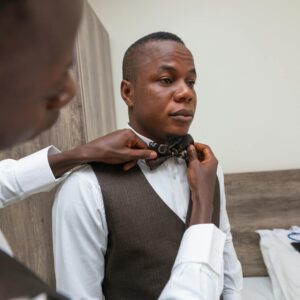 A groom getting ready for his wedding with help indoors, adjusting a bow tie.