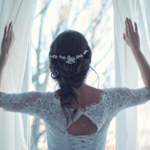 Beautiful bride in lace dress looks out a sunlit window, capturing a moment of serene elegance.