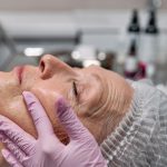 Beautician applies facial treatment on elderly woman with pink gloves in a clinic setting.